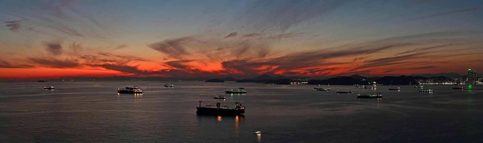 Sunset over Busan's shipping lane, cargo ships anchored offshore with the city skyline in the distance